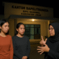 Tiga perempuan berdiri di depan kantor Bapelitbangda Kabupaten Lebak pada malam hari; dua di antaranya remaja tuna rungu sedang berkomunikasi dengan penerjemah bahasa isyarat, menggambarkan proses kesaksian dalam dugaan kasus asusila di lingkungan pemerintahan.