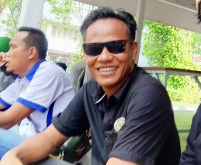 Seorang pria mengenakan kacamata hitam dan kemeja hitam terlihat berada di area publik pada siang hari.